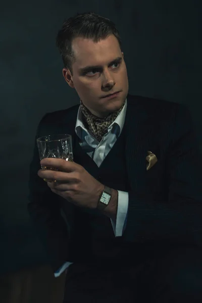 Homme de mode rétro en costume sombre tient un verre de whisky . — Photo