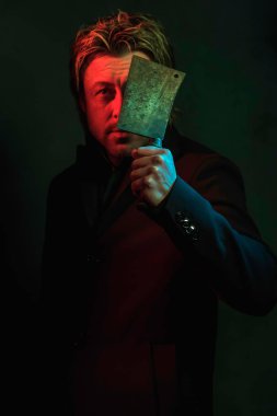 Man holding meat cleaver in front of face. Standing in red and g
