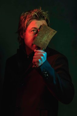 Man holding meat cleaver in front of face. Standing in red and g