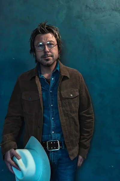 戴眼镜的男人 穿着斜纹棉布衬衫和棕色夹克 头戴牛仔帽 站在灰色的墙壁旁边 — 图库照片