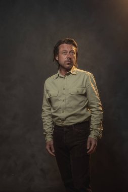 Havalı, orta yaşlı, haki gömlekli ve kahverengi pantolonlu bir adam. Alçak sesli stüdyo çekimi.