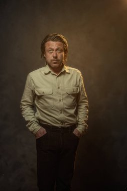 Havalı, orta yaşlı, haki gömlekli ve kahverengi pantolonlu bir adam. Alçak sesli stüdyo çekimi.