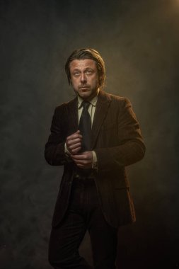 Koyu kahverengi takım elbiseli ve gri duvarın önünde siyah kravatlı havalı orta yaşlı iş adamı. Alçak sesli stüdyo çekimi.