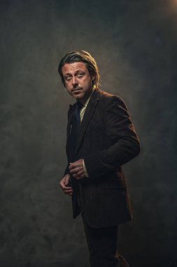 Koyu kahverengi takım elbiseli ve gri duvarın önünde siyah kravatlı havalı orta yaşlı iş adamı. Alçak sesli stüdyo çekimi.