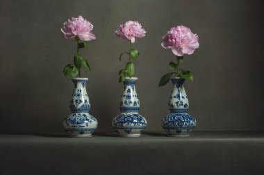 Gri bir odadaki masada pembe şakayıklı üç delft mavi vazo..