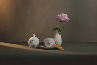 Çay fincanı, çay tabağı ve kurabiye, şeker kasesi ve şakayıklı beyaz vazo sabah güneşinde gri bir odada..