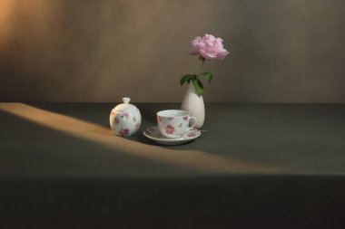 Çay fincanı çay tabağı, şeker kasesi ve şakayıklı beyaz vazo sabah güneşinde gri bir odada..