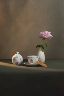 Çay fincanı, çay tabağı ve kurabiye, şeker kasesi ve şakayıklı beyaz vazo sabah güneşinde gri bir odada..