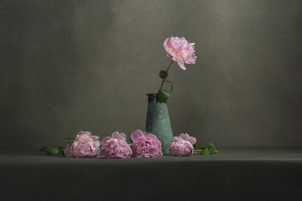 Vintage pottery vase with one peony and some peonies lying next to it on a table in a grey room.