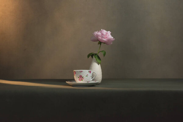 Tea cup with saucer and a white vase with peony on a table in a grey room in morning sunlight.