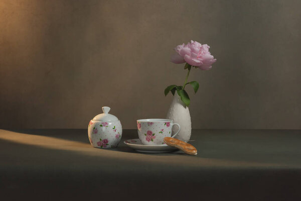 Tea cup with saucer and cookie, sugar bowl and white vase with peony on a table in a grey room in morning sunlight.