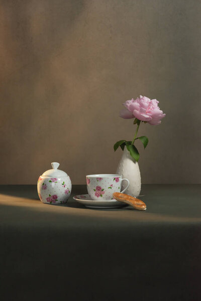 Tea cup with saucer and cookie, sugar bowl and white vase with peony on a table in a grey room in morning sunlight.