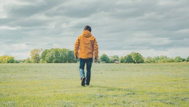 Sarı ceketli ve kot pantolonlu bir adam kırsalda çayırda yürüyor..