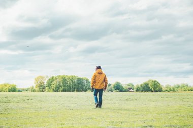 Sarı ceketli ve kot pantolonlu bir adam kırsalda çayırda yürüyor..