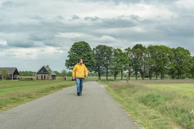 Sarı ceketli ve kot pantolonlu bir adam kırsalda yürüyüş yapıyor..
