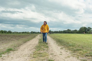 Sarı ceketli ve kot pantolonlu bir adam otlakta yürüyor..
