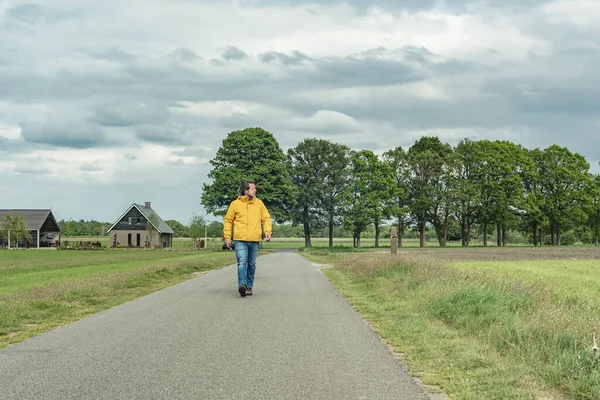 Sarı ceketli ve kot pantolonlu bir adam kırsalda yürüyüş yapıyor..