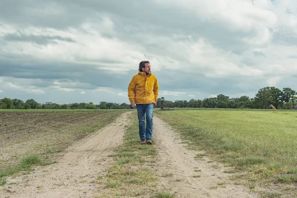 Man in yellow jacket and jeans walks on pathway in meadow.