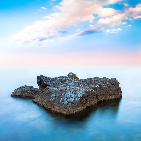 O stâncă într-un ocean albastru sub cerul înnorat la orizont. Fotografie cu expunere lungă . — Fotografie, imagine de stoc