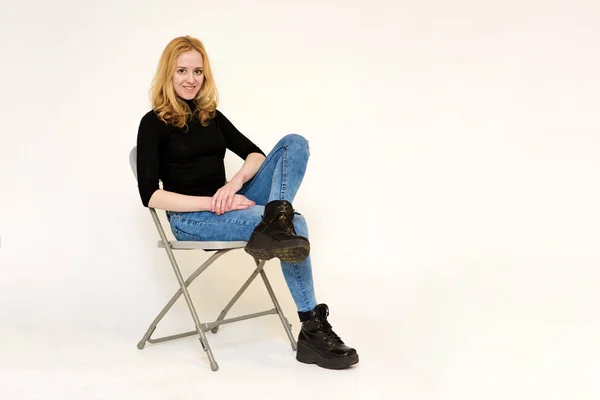 Retrato de una hermosa chica rubia en jeans sentada en una silla sobre un fondo blanco — Foto de Stock