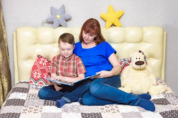 Mom and son are sitting on the bed and reading a book — Stock Photo, Image