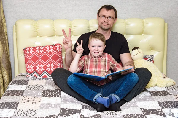 Dad and son are sitting on the bed and reading a book. — Stock Photo, Image