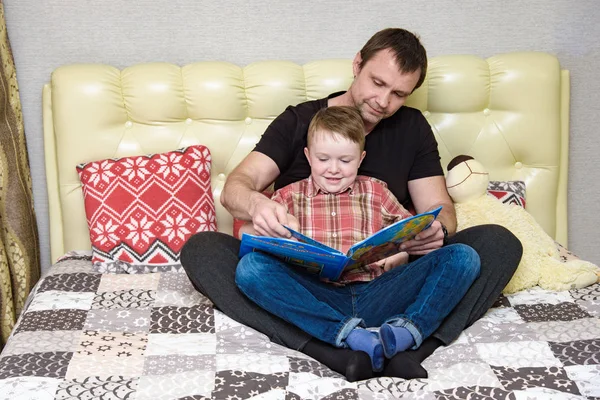 Dad and son are sitting on the bed and reading a book. — Stock Photo, Image