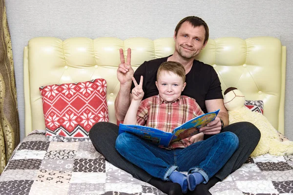 Dad and son are sitting on the bed and reading a book. — Stock Photo, Image