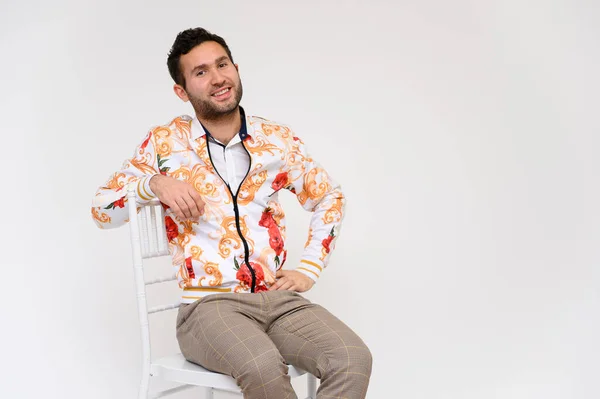 Concepto de moda para hombre. Retrato de un guapo modelo masculino, mostrando las manos, con una chaqueta blanca con un patrón floral, posando sobre un fondo blanco, menta en una silla. Pelo negro. Cerrar Studio Shot — Foto de Stock