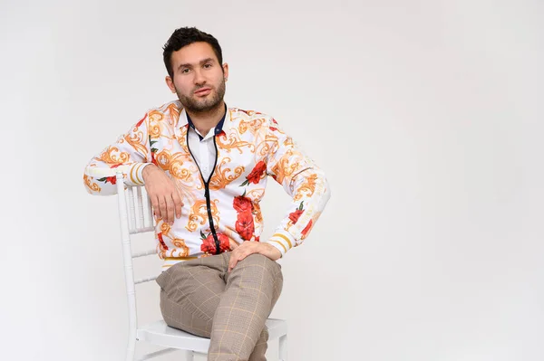 Concepto de moda para hombre. Retrato de un guapo modelo masculino, mostrando las manos, con una chaqueta blanca con un patrón floral, posando sobre un fondo blanco, menta en una silla. Pelo negro. Cerrar Studio Shot — Foto de Stock