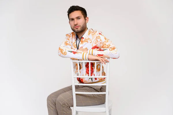 Concepto de moda para hombre. Retrato de un guapo modelo masculino, mostrando las manos, con una chaqueta blanca con un patrón floral, posando sobre un fondo blanco, menta en una silla. Pelo negro. Cerrar Studio Shot — Foto de Stock