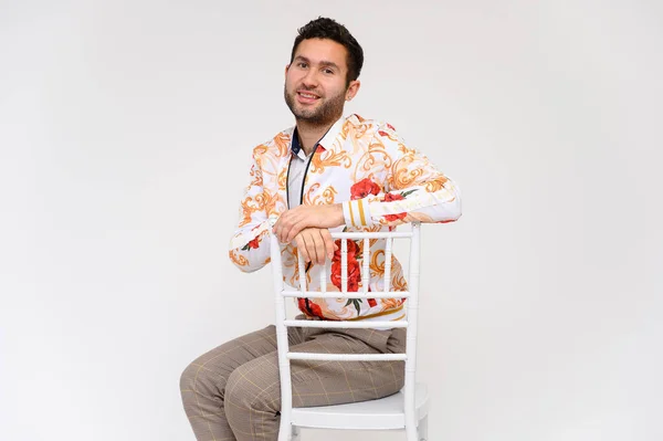 Concepto de moda masculina. Retrato de un guapo modelo masculino, mostrando las manos, con una chaqueta blanca con un patrón floral, posando sobre un fondo blanco, menta en una silla. Pelo negro. Cerrar Studio Shot — Foto de Stock