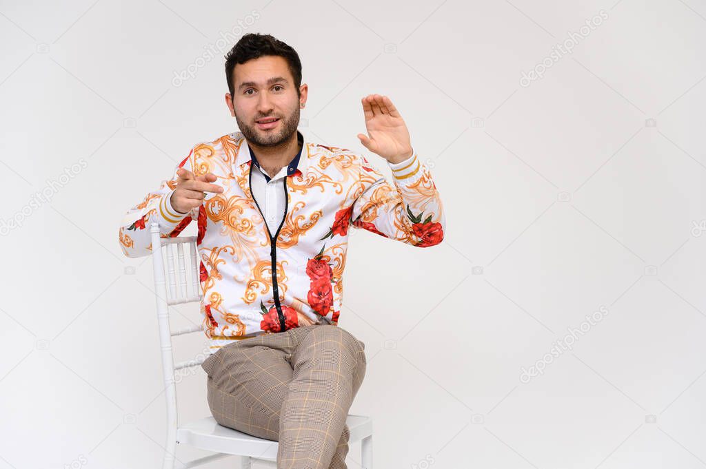 Mens fashion concept. Portrait of a handsome male model, showing hands, wearing a white jacket with a floral pattern, posing on a white background, sidmint on a chair. Black hair. Close Studio Shot