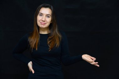 Makyajsız, temiz tenli, siyah arka planda uzun kahverengi saçlı yetişkin bir kadının portresi. Siyah bir tişörtle kameranın önünde duruyor ve ellerini gösteriyor..