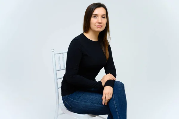 Portrait of an adult woman without makeup with clean skin, with long brown hair over her face on a white background. Sits on a chair in a black T-shirt, shows with his hands emotions. — Stock Photo, Image