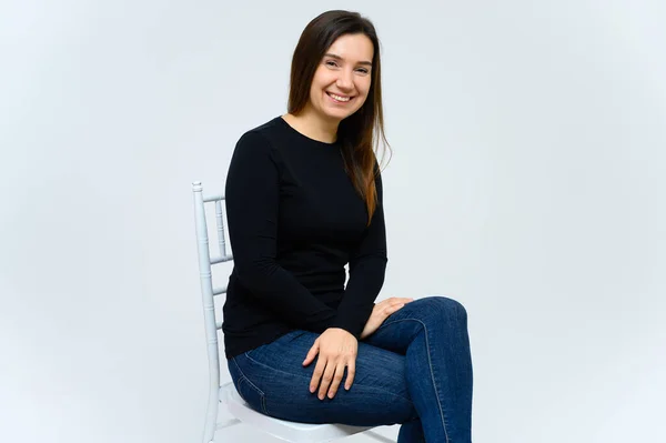 Porträt einer erwachsenen Frau ohne Make-up mit sauberer Haut, mit langen braunen Haaren vor weißem Hintergrund. sitzt im schwarzen T-Shirt auf einem Stuhl, zeigt mit den Händen Emotionen. — Stockfoto