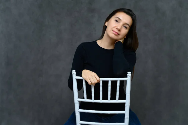 Porträtt av en vuxen kvinna utan makeup med ren hud, med långt brunt hår över ansiktet på en grå bakgrund. Sitter på en stol i en svart t-shirt, visar med händerna känslor. — Stockfoto