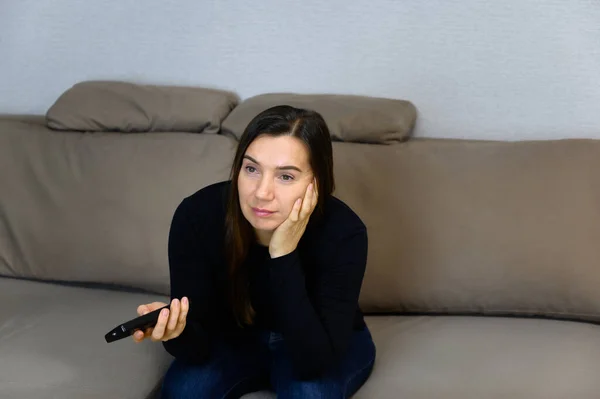 Portrait of an adult woman with long brown hair. Sitting on a sofa at home, watching TV with a remote control in his hands, showing different emotions — Zdjęcie stockowe