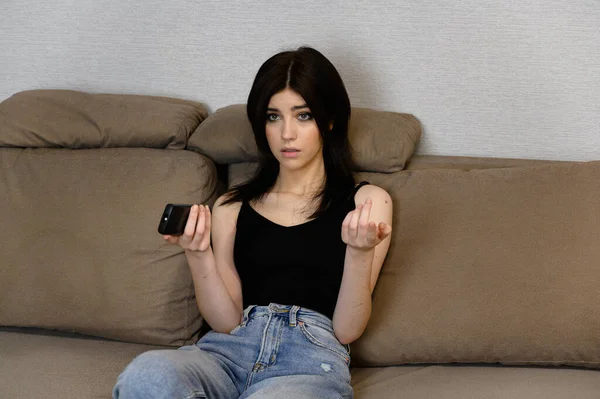 Portrait of a young girl with long black hair, clean skin of the face. Sitting on a sofa at home, watching TV with a remote control in his hands, showing different emotions — Stock Photo, Image