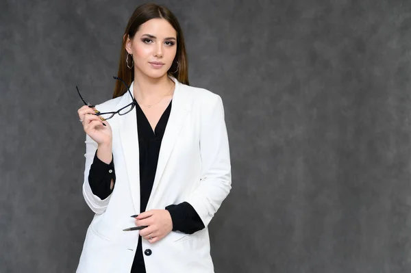 Retrato de cerca de una joven secretaria guapa con el pelo largo y negro en un traje de negocios, sobre un fondo gris. De pie justo delante de la cámara en diferentes poses con emociones . — Foto de Stock