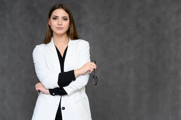 Retrato de cerca de una joven secretaria guapa con el pelo largo y negro en un traje de negocios, sobre un fondo gris. De pie justo delante de la cámara en diferentes poses con emociones . — Foto de Stock