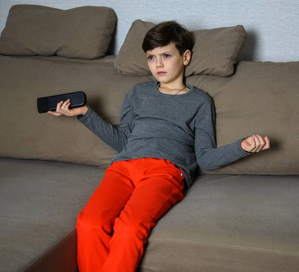 Concetto ragazzo adolescente guardando la TV a casa seduto su un divano con emozioni diverse. Foto di un bambino in una t-shirt grigia e pantaloni rossi. Si siede in pose diverse con il telecomando in mano . — Foto Stock