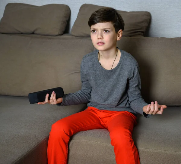 Concept boy teenager watching TV at home sitting on a sofa with different emotions. Photo of a child in a gray t-shirt and red pants. Sits in different poses with the remote control in his hands. — Stock Photo, Image