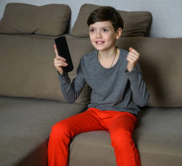 Koncepció fiú tinédzser néz TV-t otthon ül egy kanapén különböző érzelmek. Fénykép egy gyerekről szürke pólóban és piros nadrágban. Különböző pózokban ül, távirányítóval a kezében.. — Stock Fotó