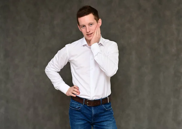 Concept of a pleasant young man manager talking to the camera. Photo Portrait of a secretary guy on a gray background in blue jeans and a white shirt in various poses. — Stock Photo, Image