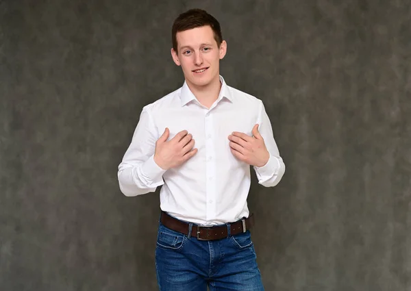 Concept of a pleasant young man manager talking to the camera. Photo Portrait of a secretary guy on a gray background in blue jeans and a white shirt in various poses. — Stock Photo, Image