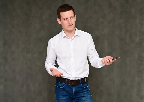 Conceito de um jovem gerente agradável conversando com a câmera. Foto Retrato de um cara secretário em um fundo cinza em jeans azul e uma camisa branca em várias poses . — Fotografia de Stock