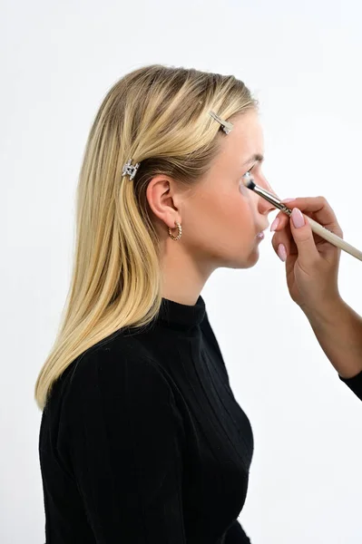 Concept de cabinet cosmétique. Brossez le maquillage avec un modèle d'un cosmétologue dans un studio cosmétique. Modèle Blond est assis sur le côté de la caméra . — Photo