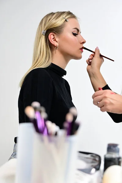 Cosmetic cabinet concept. Brush makeup with a model by a cosmetologist in a cosmetic studio. Model Blond is sitting sideways to the camera. — ストック写真