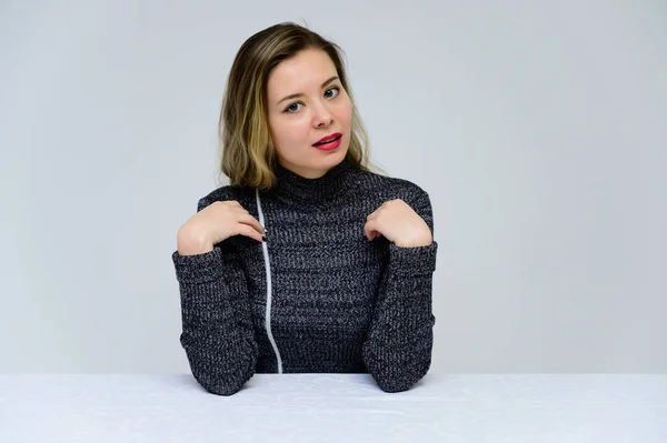 Concepto de una mujer parlante sentada en una mesa frente a la cámara. Foto de una chica bastante sonriente con el pelo rizado con emociones en un vestido gris sobre un fondo blanco . — Foto de Stock